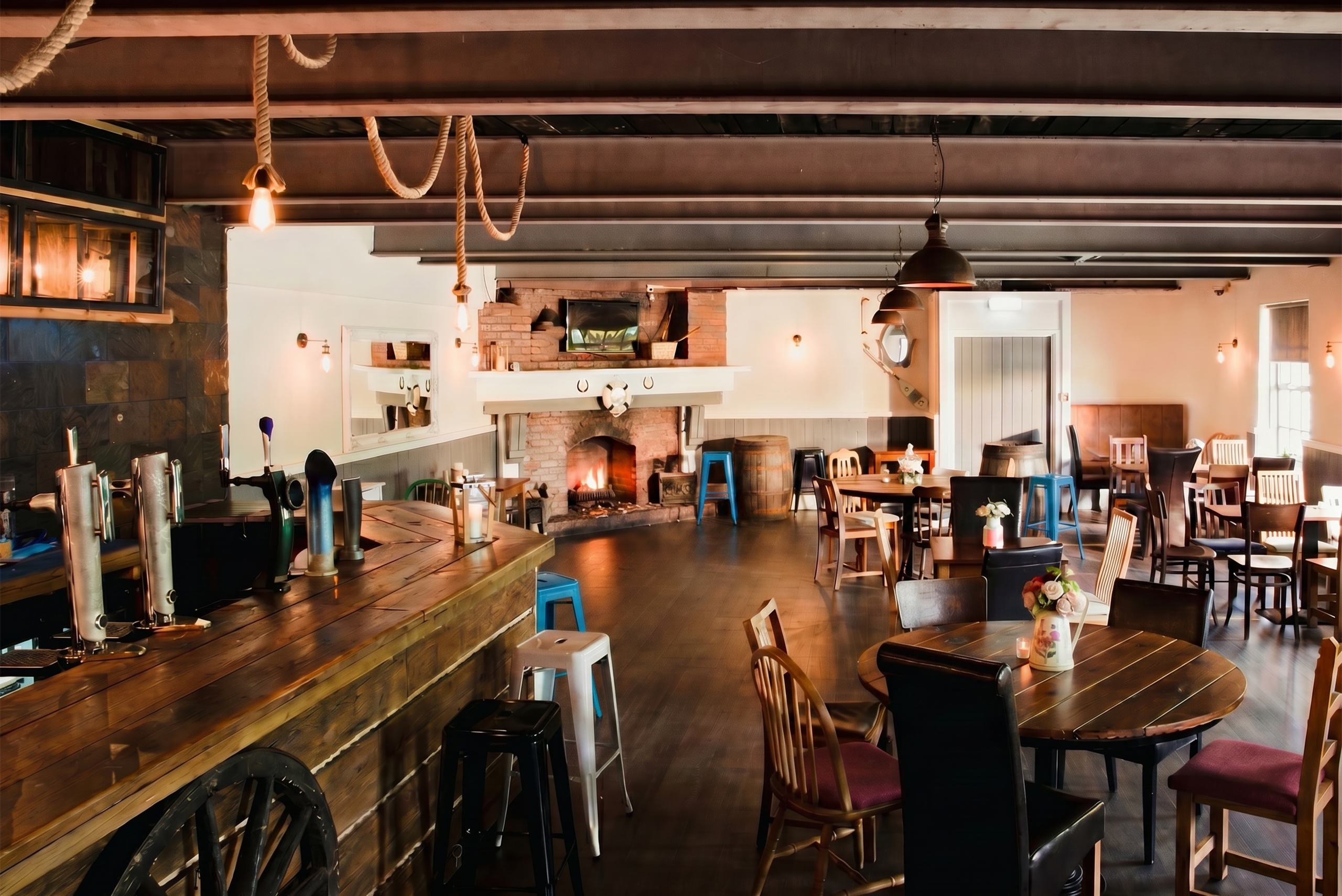 English Pub Wide interior of a rustic pub with wooden beams, hanging rope lights, and a wooden bar on the left; tables and chairs scattered throughout.